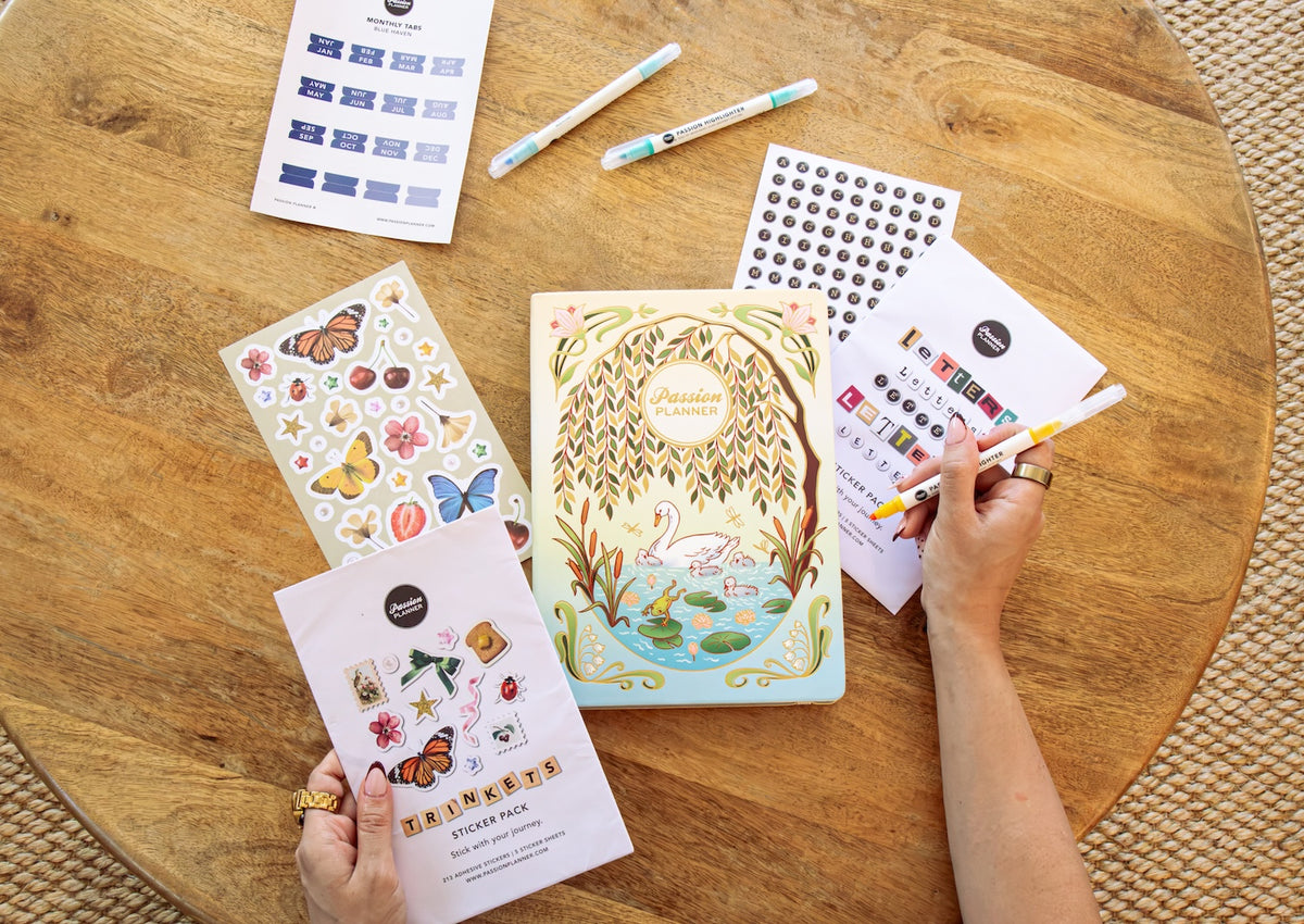 A top-down view of a person's hands holding a pastel blue and yellow illustrated 2027 planner on a round wooden table. Surrounding the planner are sticker sheets, monthly tab stickers, and highlighters.