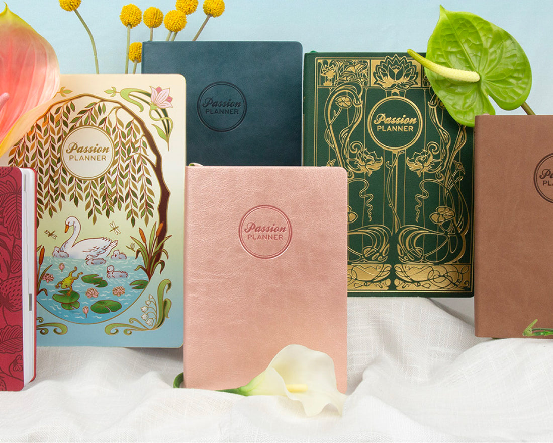 A group of Passion Planner notebooks in various colors and designs, arranged on a white fabric surface with decorative flowers and leaves against a light blue background.