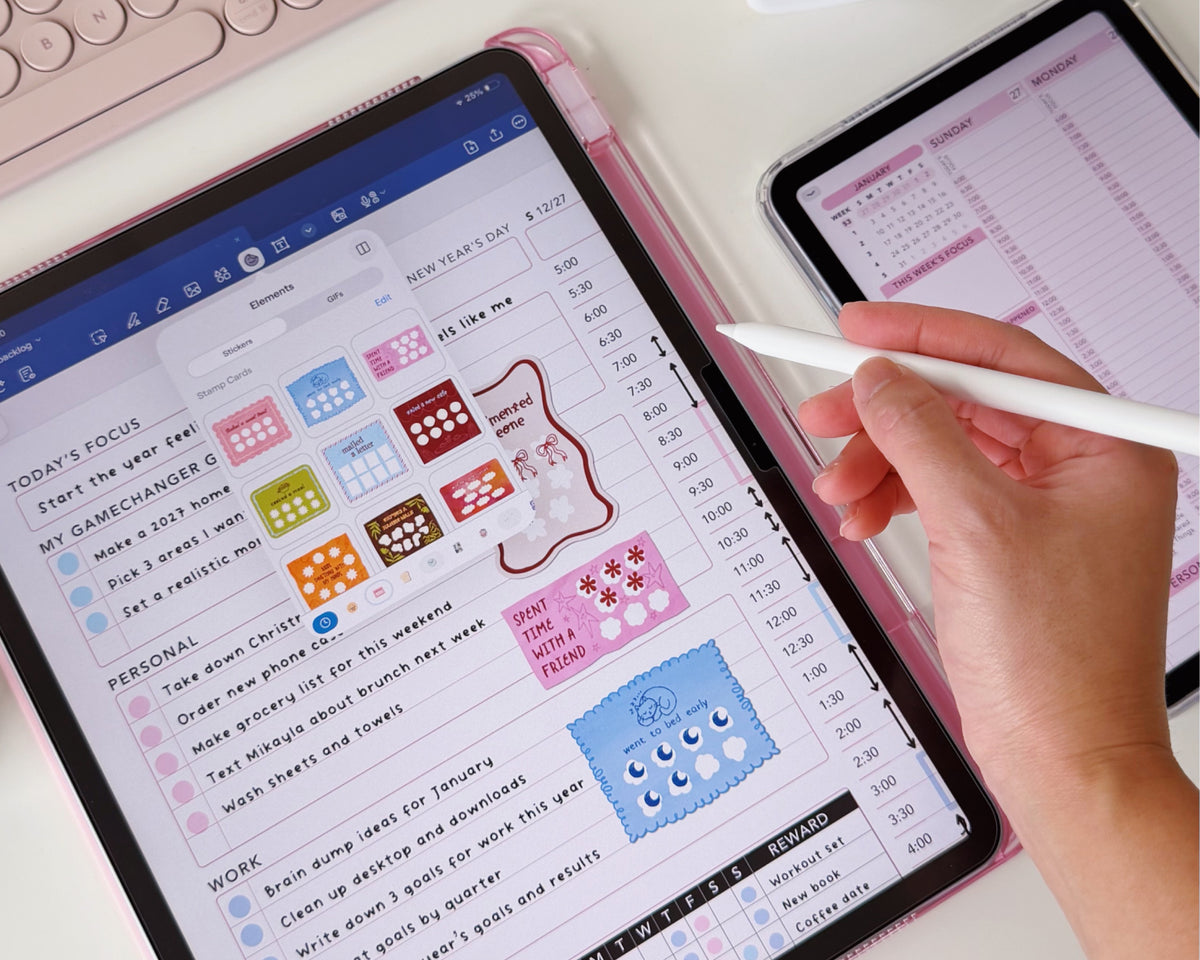 A close-up of a hand holding a white stylus over a pink tablet case displaying a colorful planner on an iPad, with another tablet partially visible on a light desk background.