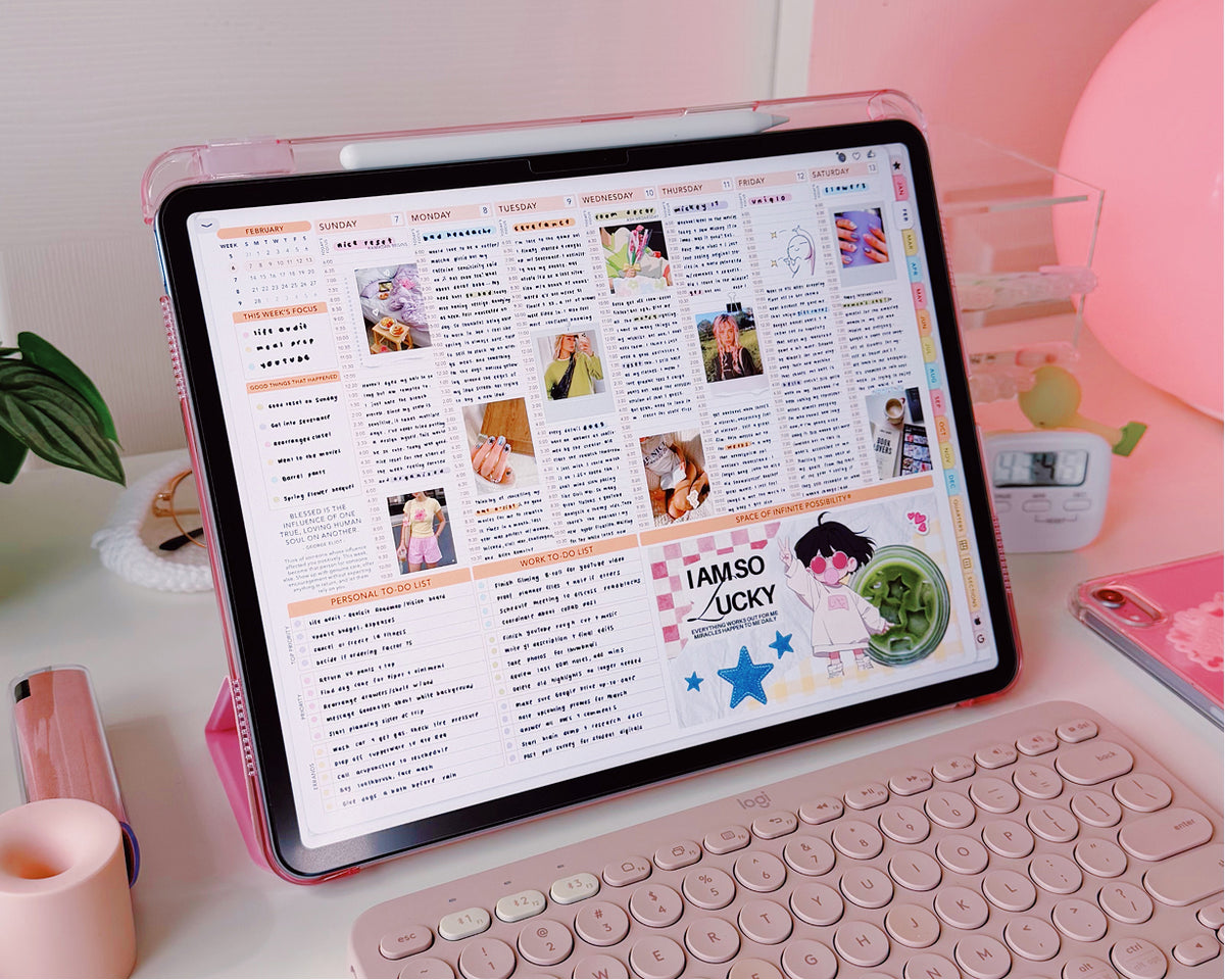 A pink tablet in a matching pink protective case and keyboard case, shown on a desk with a keyboard, planner, and pink accessories in a bright room.