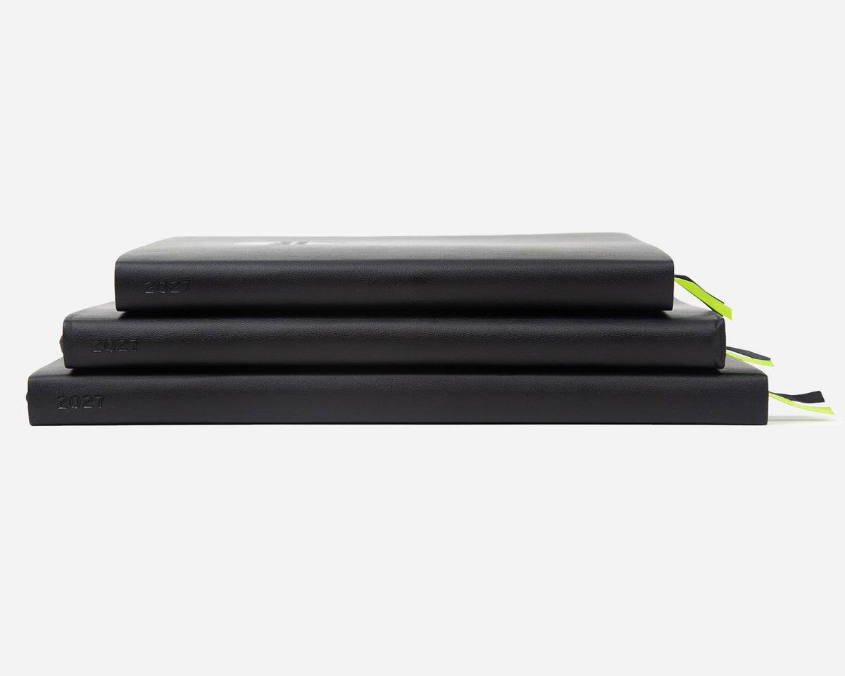 Three stacked planners in black with the year '2027' printed on the spines, and a small ribbon bookmark peeking out on the right, set against a plain white background.