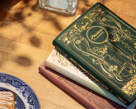 A stack of decorative 2027 planners or journals on a wooden tabletop. The top journal has a dark green cover with ornate gold floral and scrollwork and the words 'Passion Planner' visible.