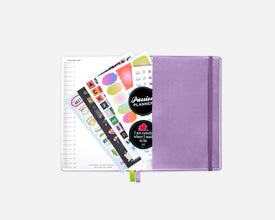 A lavender purple planner with an elastic band, shown open with several colorful decorative stickers. The image is shot on a clean, light gray background.