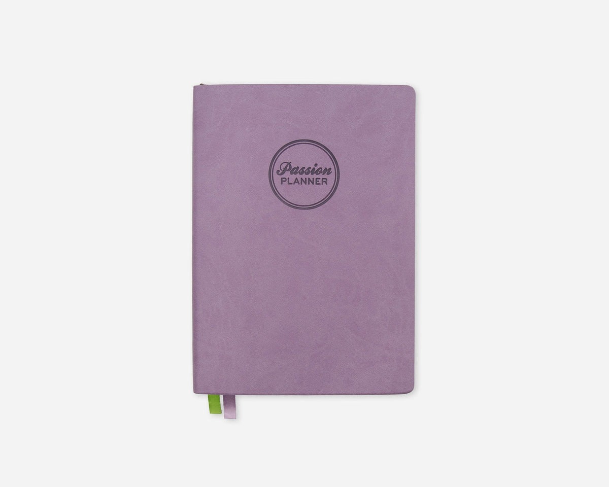A lavender purple planner with a circular debossed logo reading 'Passion Planner' on the front, shown on a clean white background with a small green and purple ribbon bookmark peeking out from the bottom.