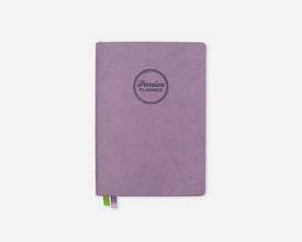 A lavender purple planner with a circular debossed logo reading 'Passion Planner' on the front, shown on a clean white background with a small green and purple ribbon bookmark peeking out from the bottom.
