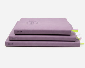 Three 2027 lavender colored planners stacked on a light gray background. 