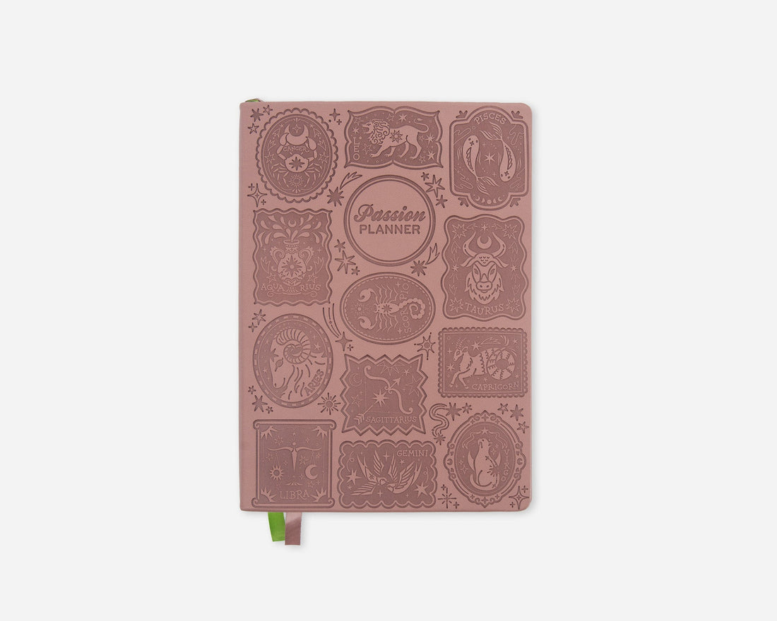 A dusty rose pink 2027 planner with embossed zodiac animal illustrations and a small green ribbon bookmark, shown on a light gray background.