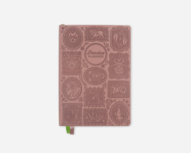 A dusty rose pink 2027 planner with embossed zodiac animal illustrations and a small green ribbon bookmark, shown on a light gray background.