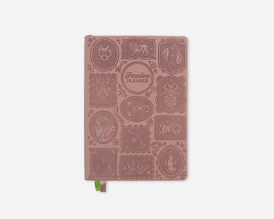 A dusty rose pink 2027 planner with embossed zodiac animal illustrations and a small green ribbon bookmark, shown on a light gray background.