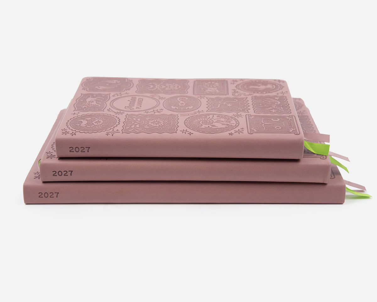 Three 2027 mauve colored planners stacked on a light gray background. 