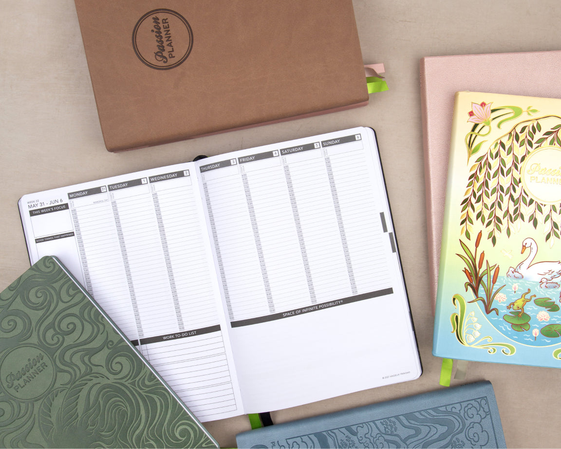 A styled flat-lay of several planners and notebooks on a beige surface. The main subject is an open white weekly planner with black lines and gray headers, surrounded by closed planners in green, brown kraft paper, pink, and blue, all branded with 'Passion Planner'.