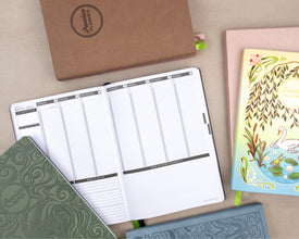 A styled flat-lay of several planners and notebooks on a beige surface. The main subject is an open white weekly planner with black lines and gray headers, surrounded by closed planners in green, brown kraft paper, pink, and blue, all branded with 'Passion Planner'.