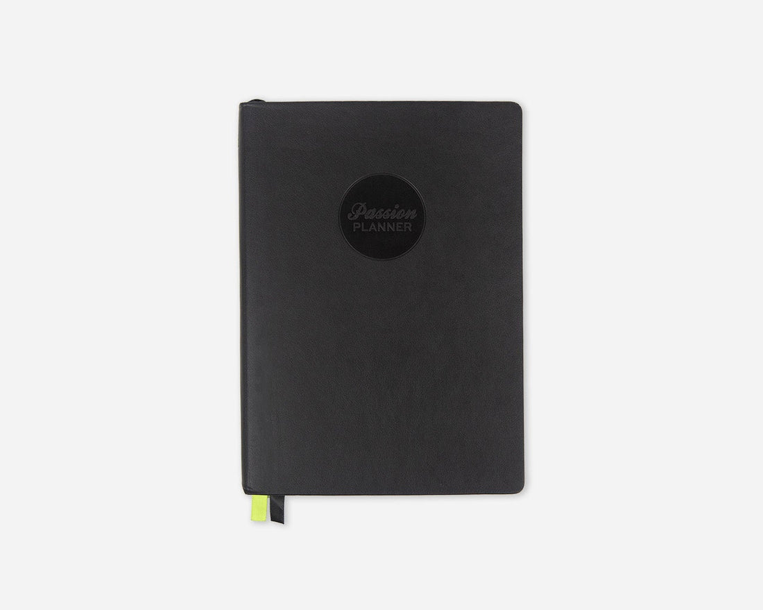 A black faux leather 2027 Passion Planner with a circular black logo on the front and a small lime green ribbon bookmark peeking out from the bottom, set against a plain light gray background.