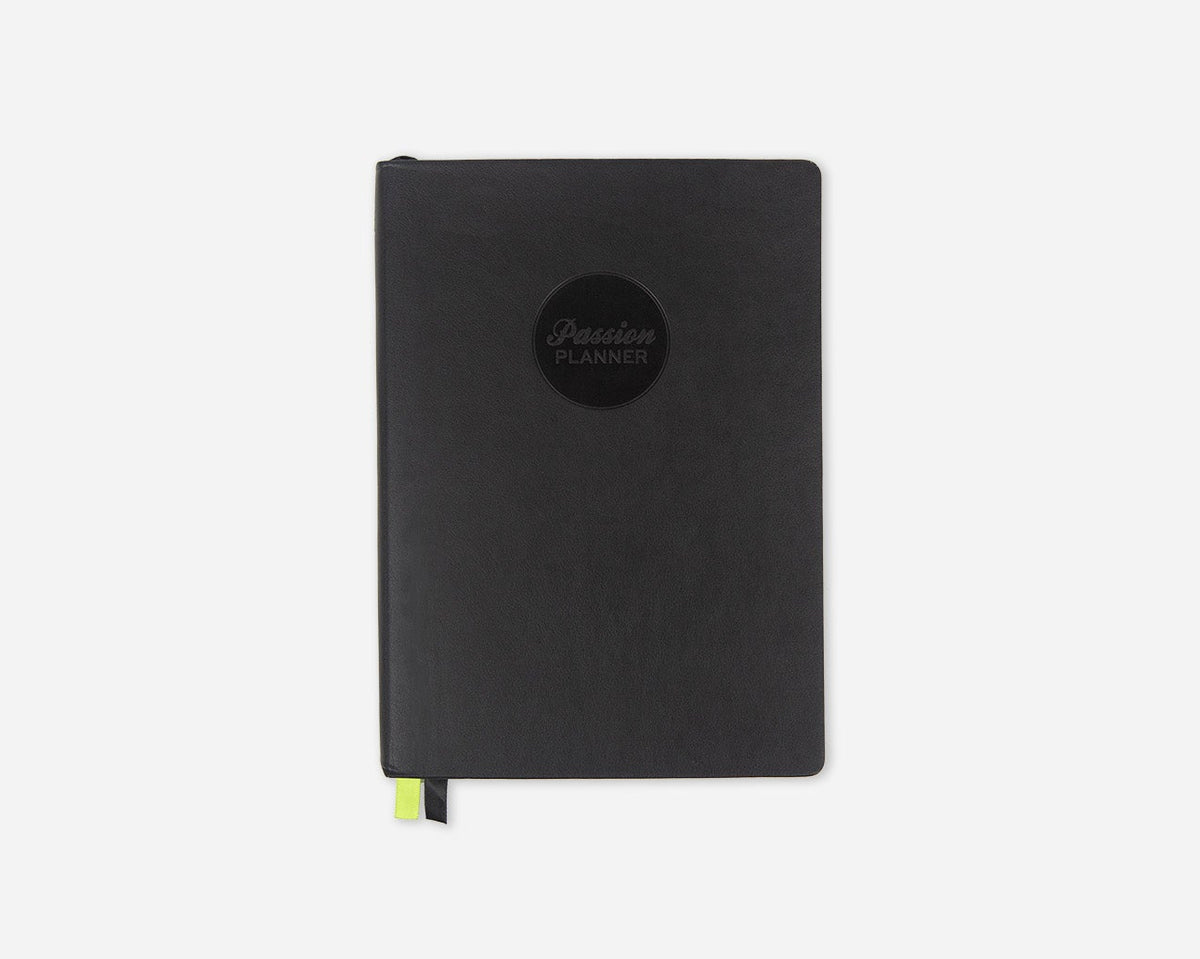 A black faux leather 2027 Passion Planner with a circular black logo on the front and a small lime green ribbon bookmark peeking out from the bottom, set against a plain light gray background.