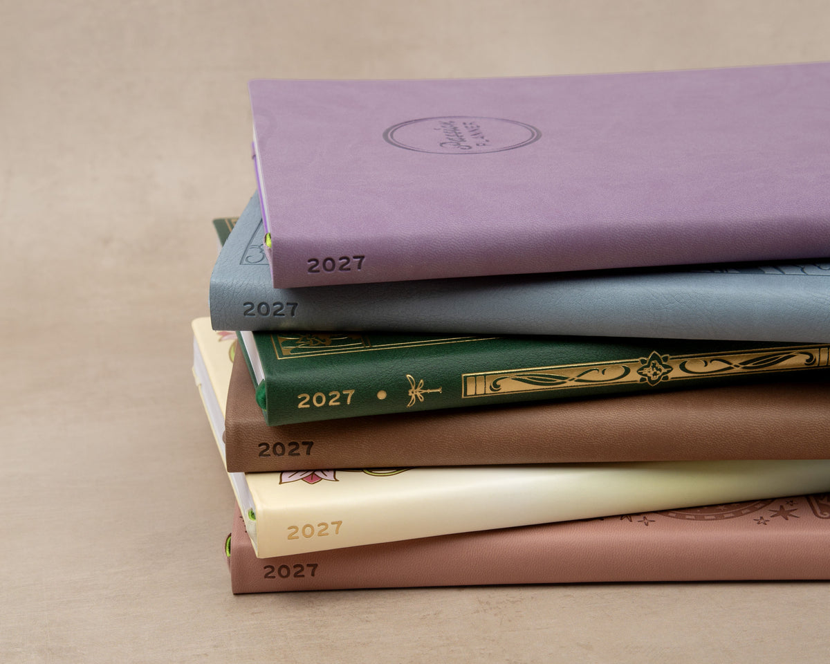 A stack of six planners on a light beige surface, each with the year 2027 visible on the cover in different muted colors including lavender, light blue, green, brown, and cream.