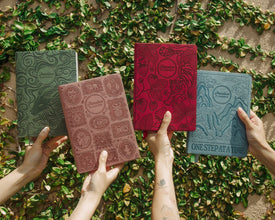 Four embossed leather planners in green, dusty rose, red, and slate blue are held up by multiple hands against a background of green ivy on a stone wall. The covers feature various embossed illustrations and phrases like 'One Step At A Time'.