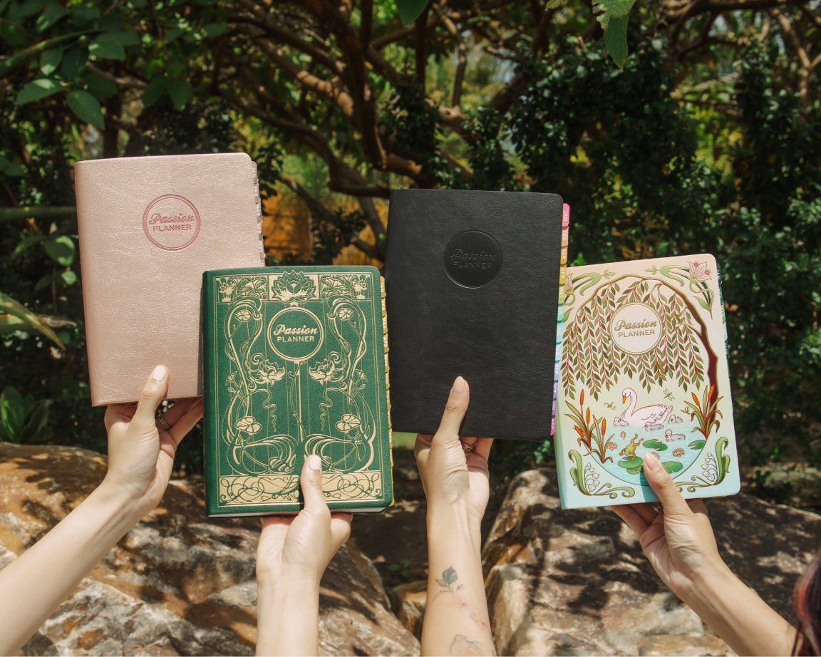 Four illustrated planners held up by multiple hands outdoors in front of a forest background. The notebooks feature pastel pink, green with gold detailing, black, and a multicolor floral design, all with the brand name 'Passion Planner' visible on the covers.