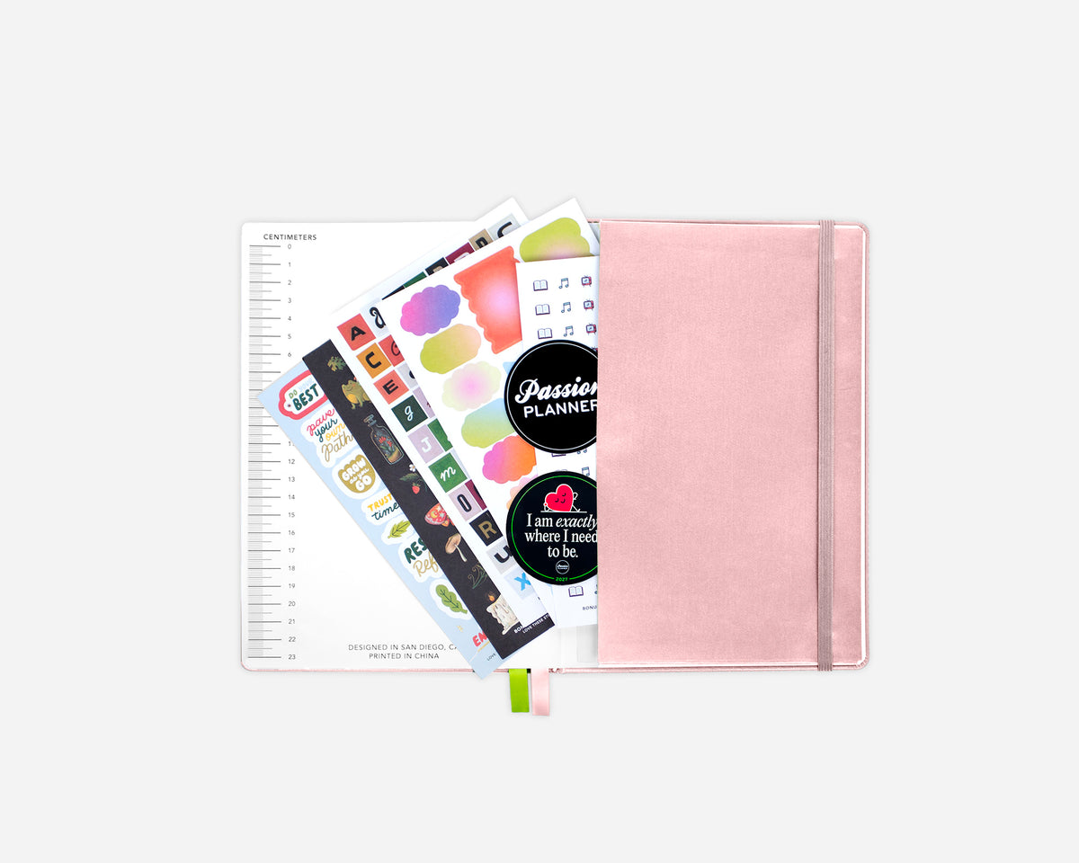 An open rose gold 2027 planner with an elastic closure, shown open with colorful sticker sheets fanned out against a light gray background.