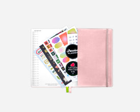 An open rose gold 2027 planner with an elastic closure, shown open with colorful sticker sheets fanned out against a light gray background.
