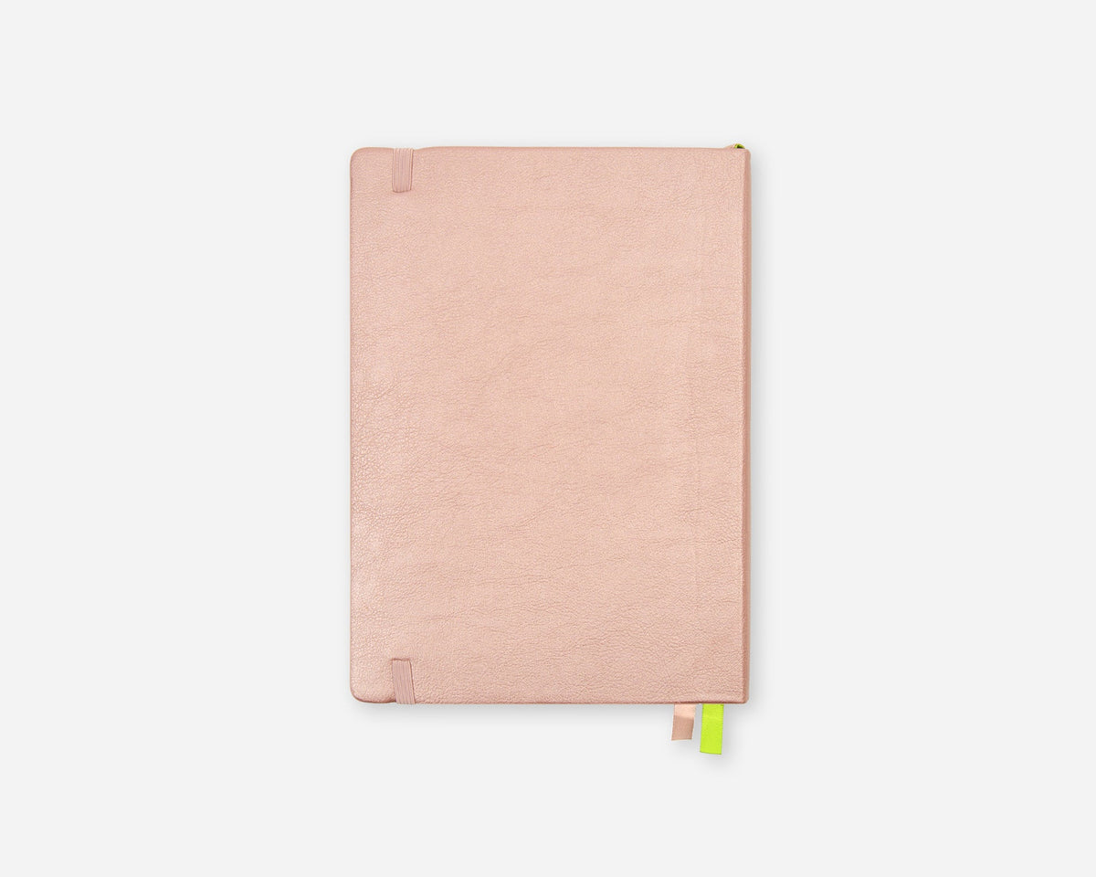 A slim, rose gold notebook with a textured cover, featuring a small green and pink ribbon bookmark hanging from the bottom edge, set against a solid light gray background.