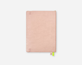 A slim, rose gold notebook with a textured cover, featuring a small green and pink ribbon bookmark hanging from the bottom edge, set against a solid light gray background.