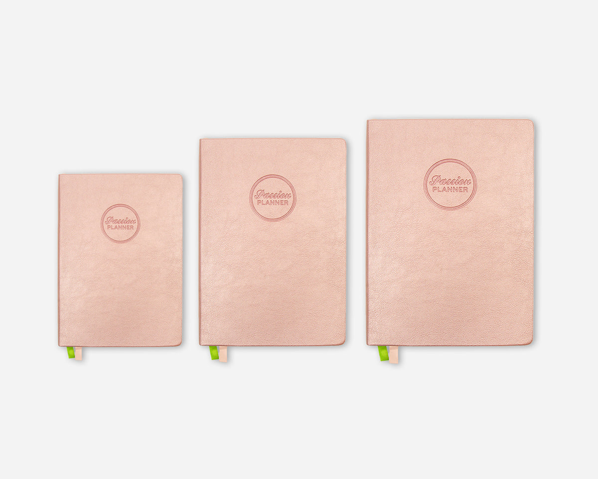 Three rose gold planner notebooks in different sizes lined up horizontally on a light gray background. Each notebook features a circular embossed logo that reads 'Passion Planner' and has a small green ribbon bookmark peeking out from the bottom.