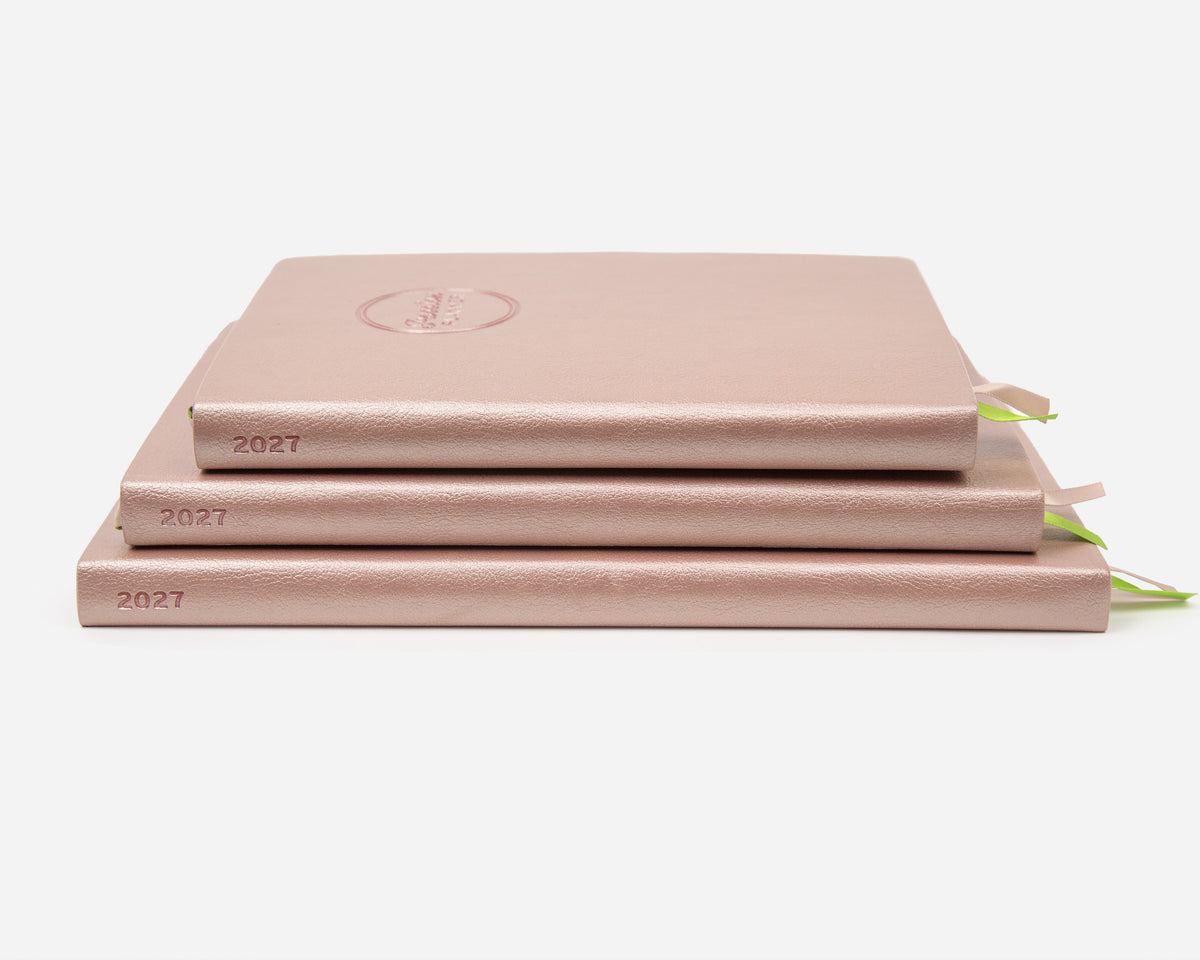 Three 2027 rose gold colored planners stacked on a light gray background. 