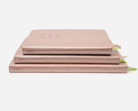 Three 2027 rose gold colored planners stacked on a light gray background. 