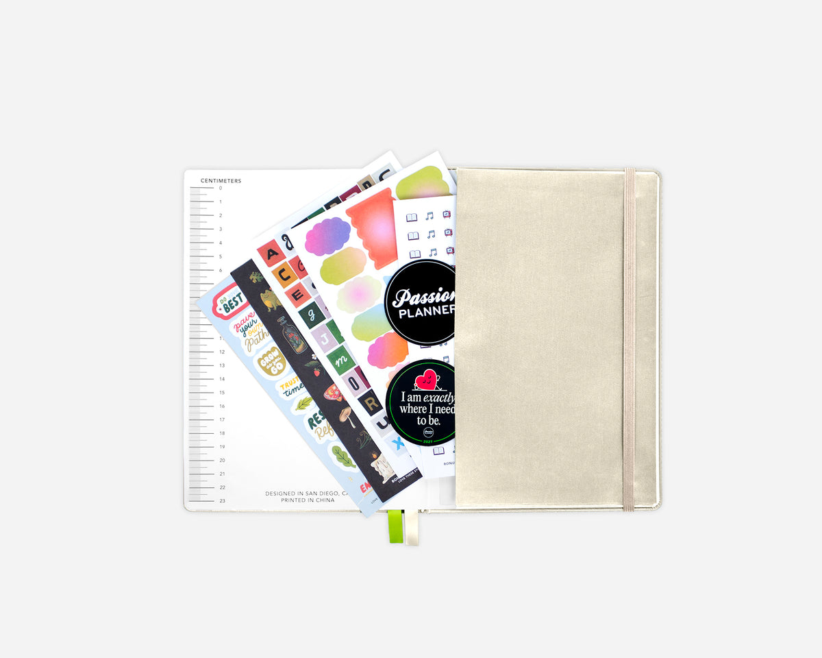 A cream-colored fabric planner sleeve with an elastic closure, shown with colorful decorative stickers. The planner features a black circular sticker with the text 'Passion Planner' and a small heart graphic.