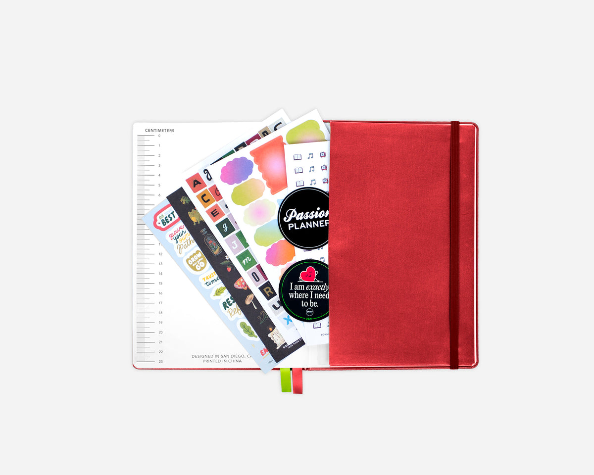 A red planner with an elastic closure, shown open with printed pages and colorful sticker sheets inside, on a light gray background.