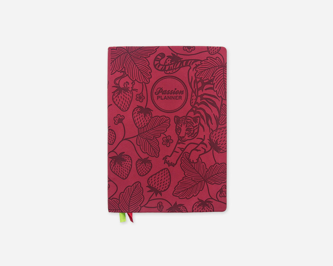 A faux leather planner with a dark red cover featuring botanical illustrations of strawberries, leaves, and a tiger, with a small green and red ribbon bookmark hanging from the bottom.