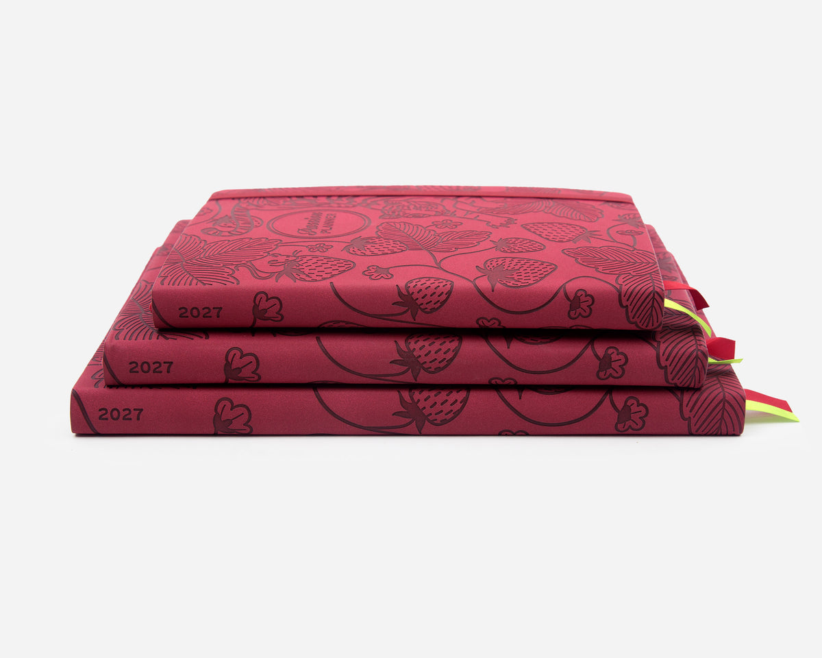 Three stacked planners with deep red covers featuring a black botanical strawberry pattern, the year '2027' printed on the spines, and small ribbon bookmarks peeking out on the right, set against a plain white background.