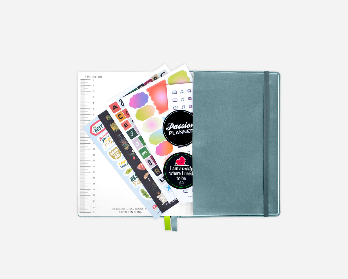 A closed blue planner with an elastic closure, shown open with sticker sheets and a lined page fanned out against a light gray background.