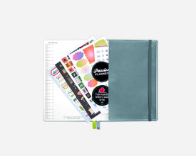 A closed blue planner with an elastic closure, shown open with sticker sheets and a lined page fanned out against a light gray background.