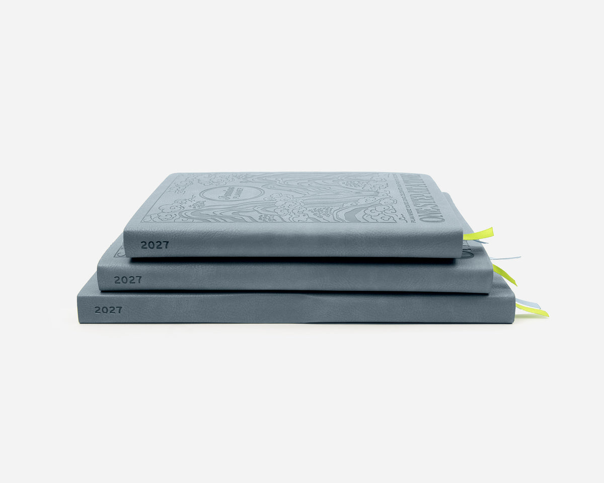 Three stacked planners in a muted blue-gray color, each embossed with the year 2027. The top journal features an ornate crest-like embossing on its cover, and small bright green ribbon bookmarks are visible on the right side.