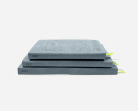 Three stacked planners in a muted blue-gray color, each embossed with the year 2027. The top journal features an ornate crest-like embossing on its cover, and small bright green ribbon bookmarks are visible on the right side.