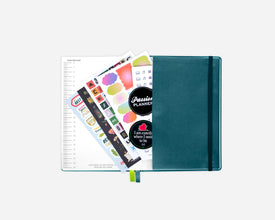 A closed teal blue planner with an elastic closure, shown open with sticker sheets and a lined page fanned out against a light gray background.
