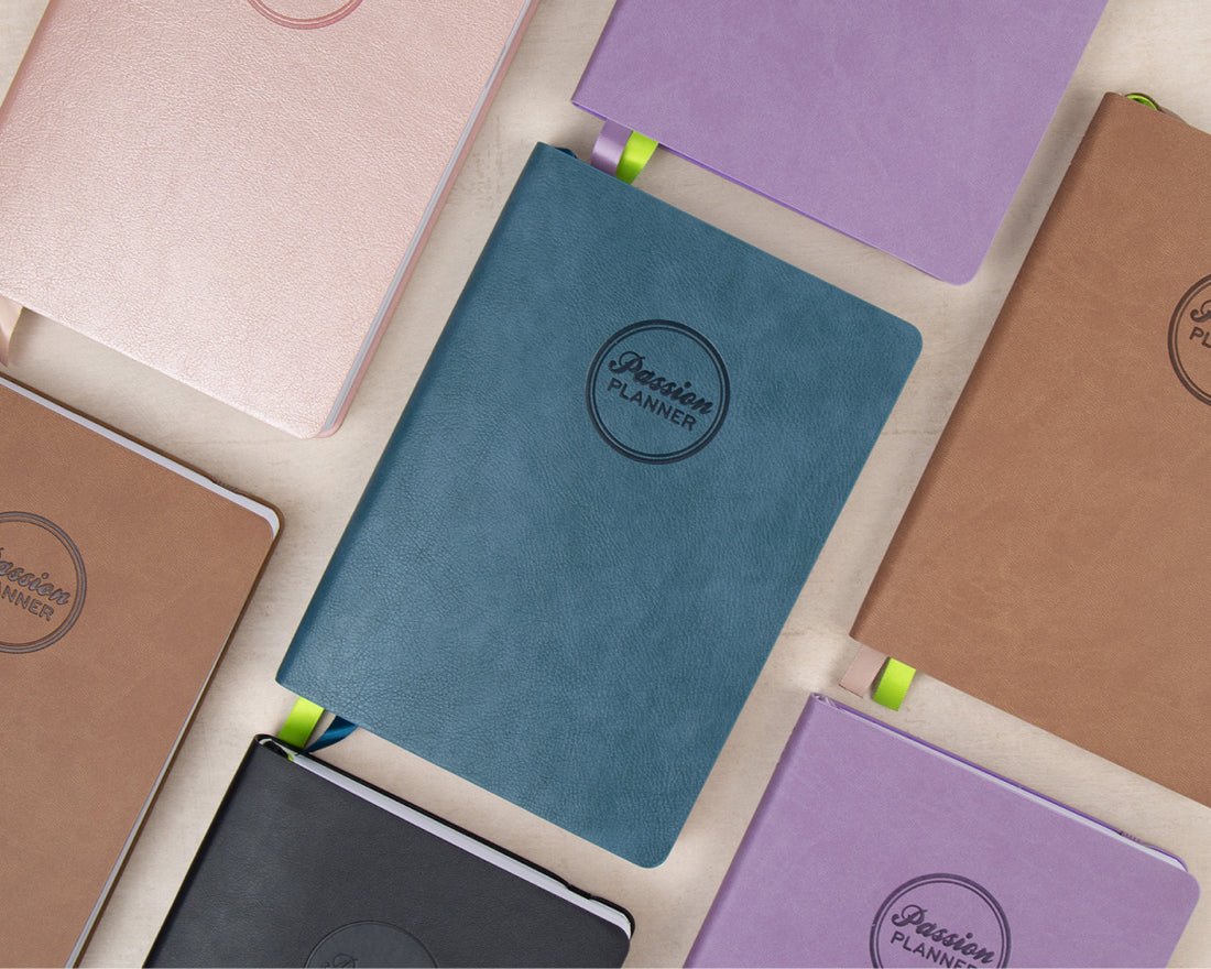 A collection of 2027 planners with textured covers in various colors, including teal, lavender, brown, pink, black, and beige, arranged in a flat-lay pattern on a light cream background. Each notebook features a circular embossed logo that reads &