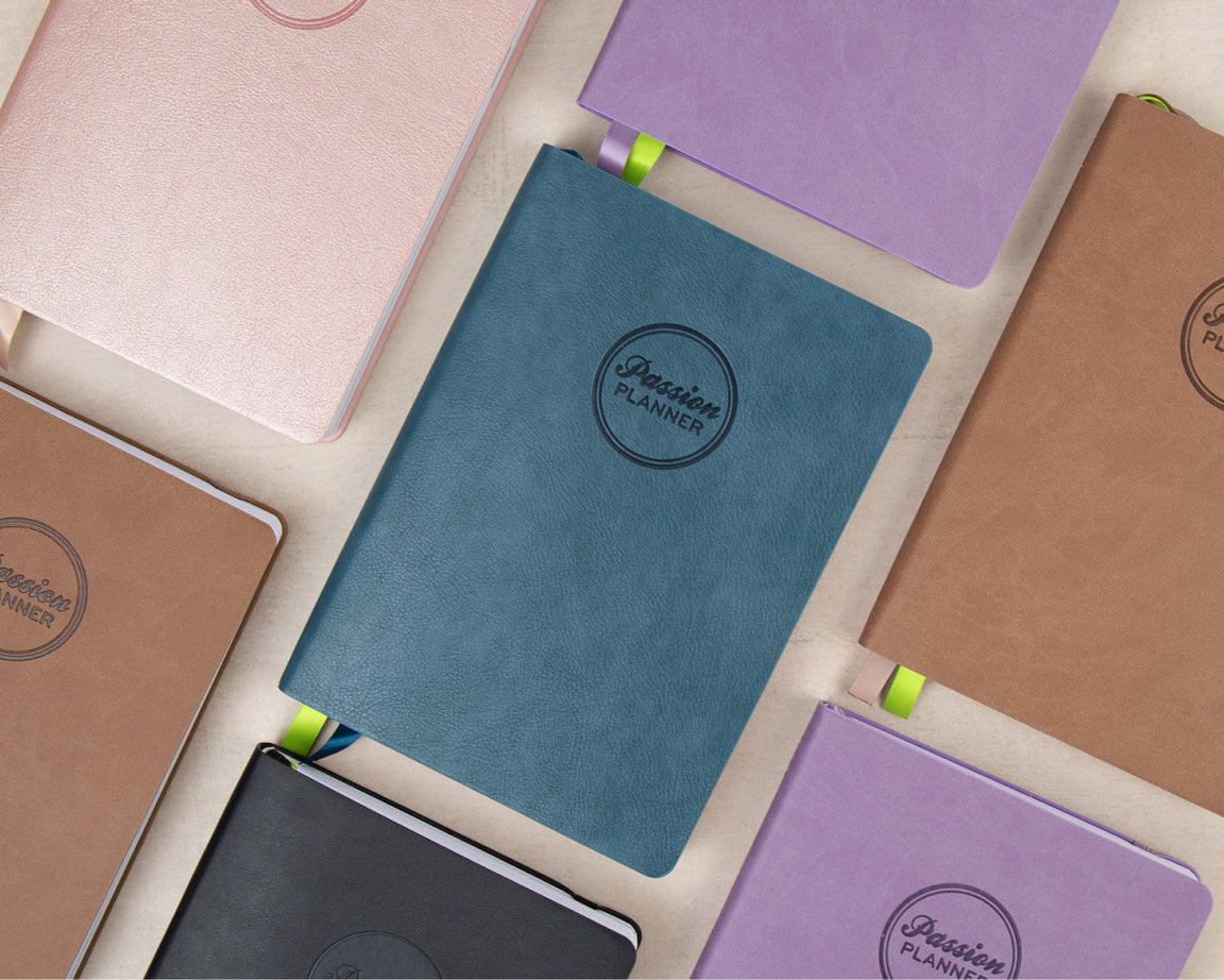 A collection of 2027 planners with textured covers in various colors, including teal, lavender, brown, pink, black, and beige, arranged in a flat-lay pattern on a light cream background. Each notebook features a circular embossed logo that reads 'Passion Planner'.