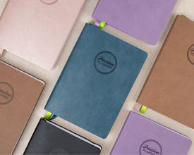 A collection of 2027 planners with textured covers in various colors, including teal, lavender, brown, pink, black, and beige, arranged in a flat-lay pattern on a light cream background. Each notebook features a circular embossed logo that reads 'Passion Planner'.