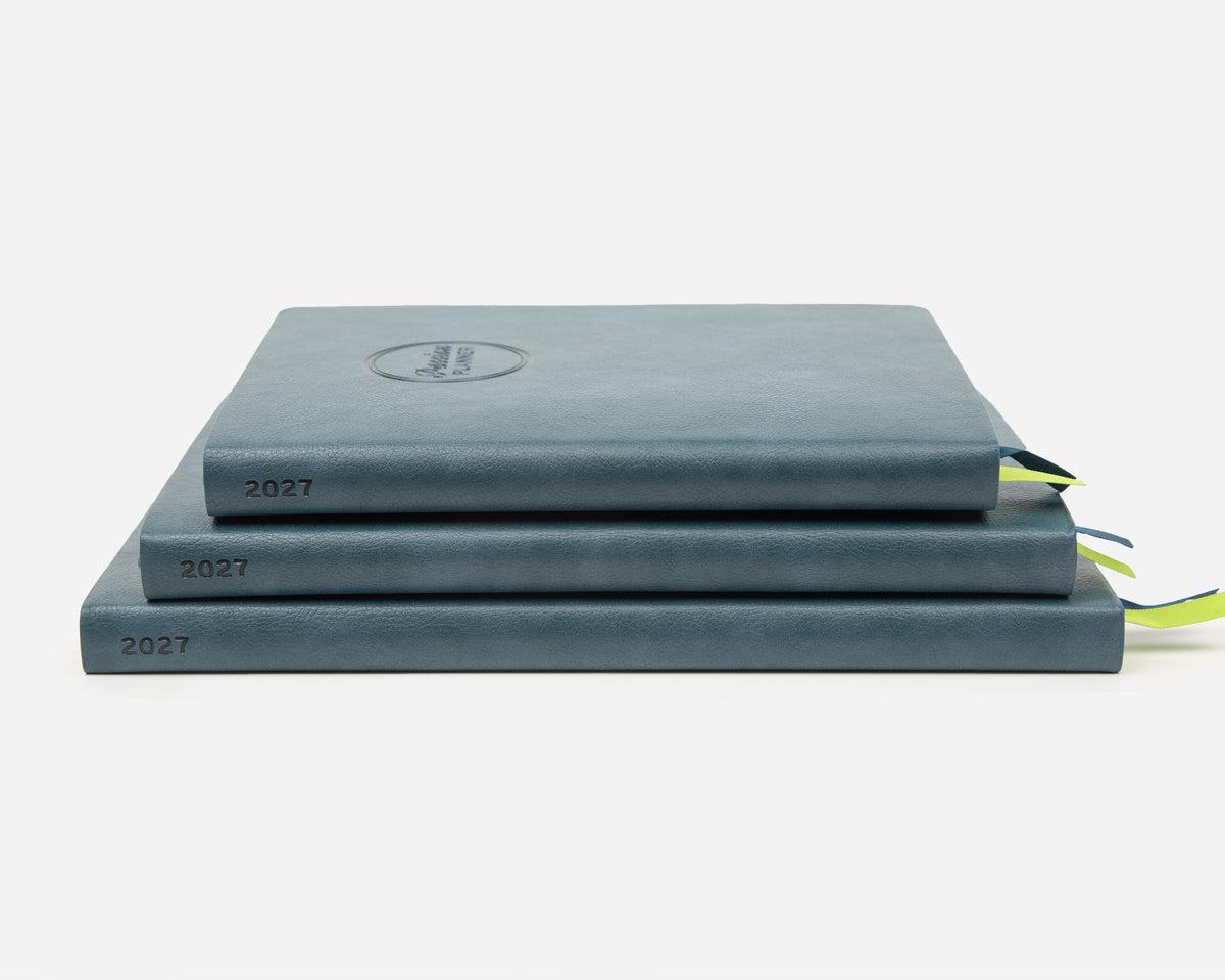 Three stacked faux leather planners in a muted slate blue-gray color, each labeled '2027' on the spine, with small ribbon bookmarks peeking out on a clean white background.