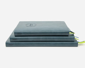 Three stacked faux leather planners in a muted slate blue-gray color, each labeled '2027' on the spine, with small ribbon bookmarks peeking out on a clean white background.