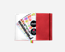 A closed red planner with an elastic closure, shown open with sticker sheets and a lined page fanned out against a light gray background.