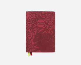 A red 2027 planner with a zodiac-themed design featuring a goat head profile and swirling patterns, with a 'Passion Planner' logo. A small ribbon bookmark with red, yellow, and green tabs is visible at the bottom.