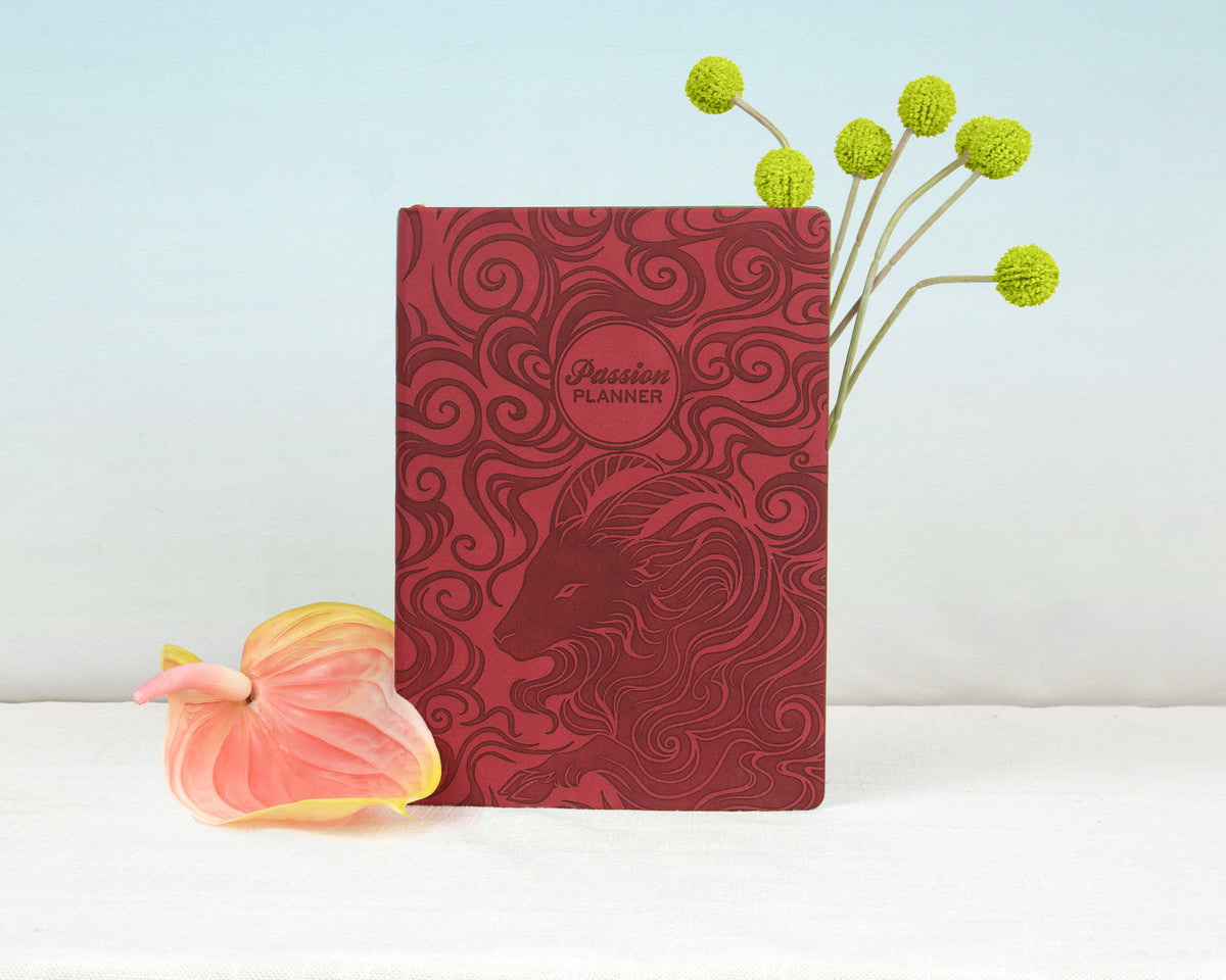 A red embossed planner with a ram design and the words 'Passion Planner' on the cover, styled on a white surface with a pink flower and green stems against a light blue background.