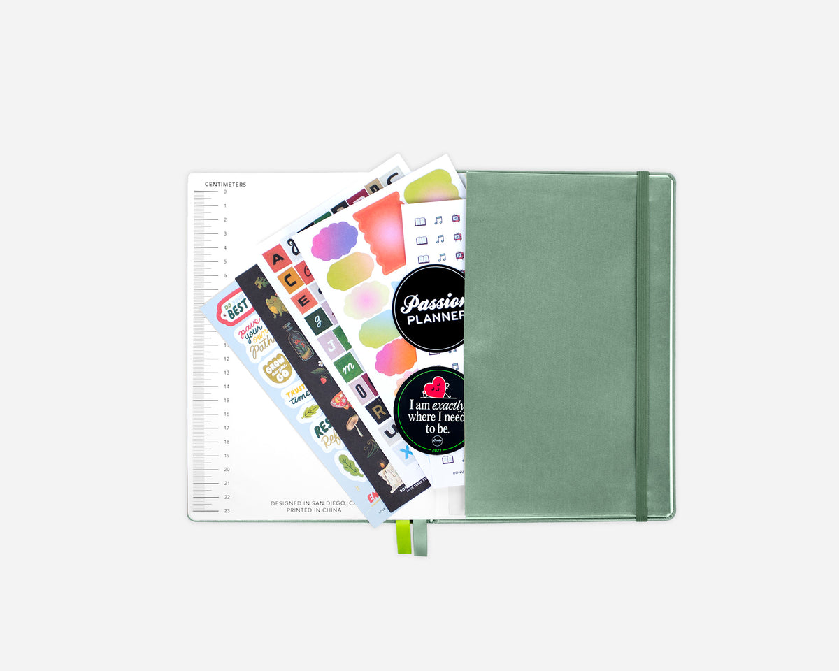 A closed sage green planner with an elastic closure, shown open with sticker sheets and a lined page fanned out against a light gray background.