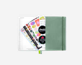 A closed sage green planner with an elastic closure, shown open with sticker sheets and a lined page fanned out against a light gray background.