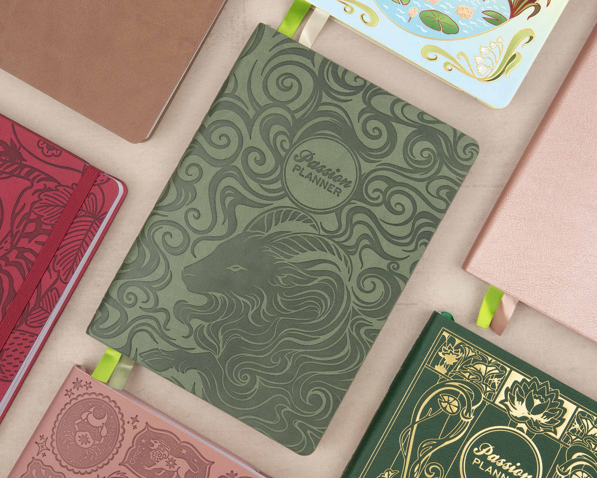 A green embossed 'Passion Planner' notebook with a goat graphic and swirl patterns, styled in a flat-lay arrangement with other colorful planners on a beige surface.