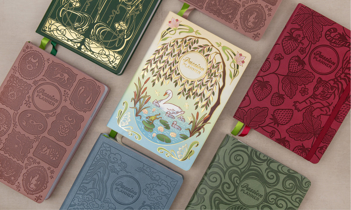 Six decorative planner  covers arranged in a flat lay on a light neutral background. The covers feature embossed floral and decorative patterns in muted tones including dusty rose, forest green, coral red, light blue, and sage green, with one central cover showing a whimsical tree-of-life illustration with a swan and pond.