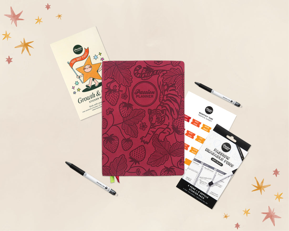 A styled flat-lay of stationery items on a light beige background with small star illustrations. The main product is a red planner with botanical strawberry and leaf patterns, labeled &